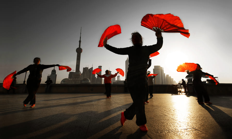Personas practicando la danza tradicional con abanicos rojos al amanecer en Shanghái, con la Torre de la Perla Oriental de fondo, representando las experiencias auténticas y viajes culturales a China.